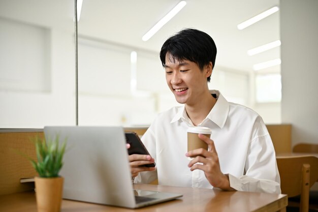 L'uomo asiatico felice si siede in una caffetteria sorseggiando caffè e usando il suo smartphone