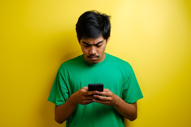 L'uomo asiatico gioca al gioco mobile sul suo smartphone con un'espressione seria o arrabbiata su sfondo giallo