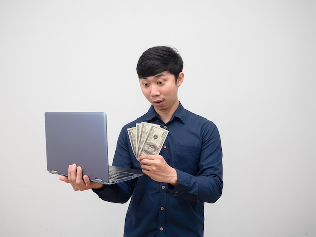 L'uomo asiatico guarda i soldi in mano e tiene in mano il laptop sentendosi stupito su sfondo bianco