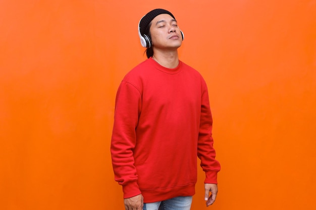 L'uomo asiatico in cuffia wireless chiude gli occhi e si gode la canzone mentre si trova in piedi su sfondo arancione