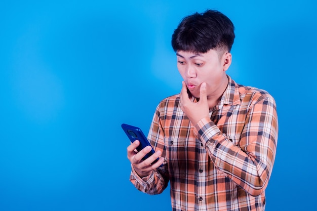 L'uomo asiatico sembra sorpreso dalle buone notizie che ha ricevuto dal suo smartphone. Gli uomini mostrano movimenti scioccati con gli occhi sporgenti mentre sono sullo smartphone su sfondo blu