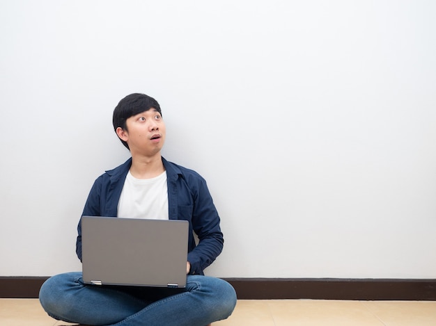 L'uomo asiatico si siede sul muro bianco del pavimento con il laptop sentendosi eccitato wow guardando lo spazio