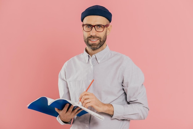 L'uomo attraente contento con stoppia indossa occhiali trasparenti cappello e camicia alla moda scrive con la penna nel blocco note si prepara a parlare davanti all'auditorium isolato su sfondo rosa