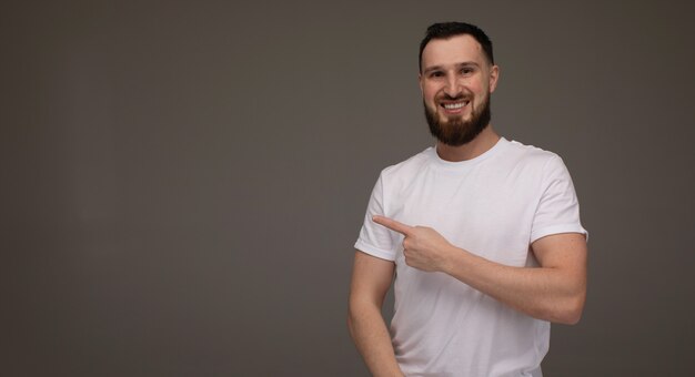 L'uomo barbuto bello in abbigliamento casual sta indicando via, sta esaminando la macchina fotografica e sta sorridendo, su fondo grigio