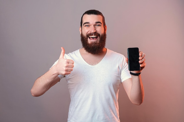 L'uomo barbuto bello sta mostrando lo schermo di un telefono e un mi piace per l'approvazione