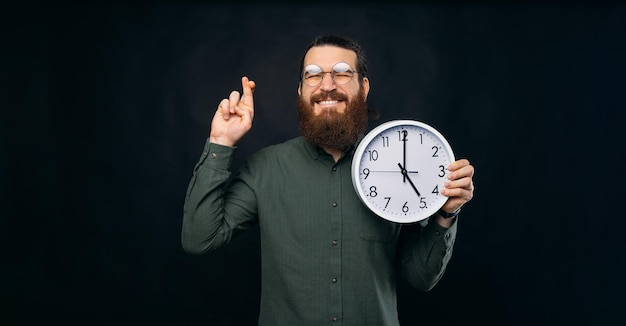 L'uomo barbuto con gli occhiali sta incrociando le dita per un desiderio mentre tiene in mano un orologio bianco su sfondo nero