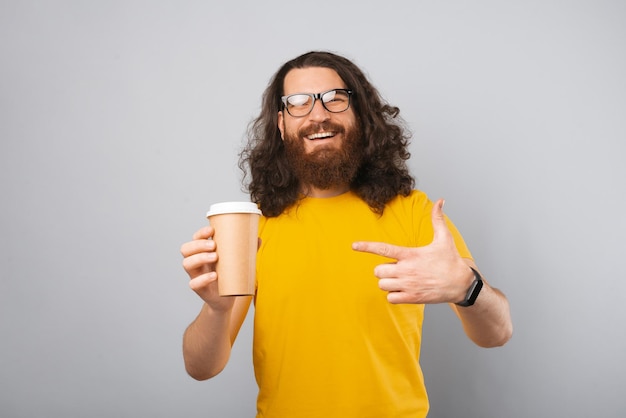 L'uomo barbuto con i capelli ondulati sta puntando a una tazza di carta da caffè per andare Studio girato su sfondo grigio