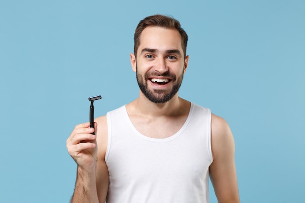 L'uomo barbuto di 20 anni in camicia bianca tiene un rasoio di plastica monouso isolato su sfondo blu pastello ritratto in studio. Concetto di procedure cosmetiche sanitarie per la cura della pelle. Simula lo spazio della copia.