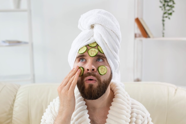 L'uomo barbuto e divertente si diverte con una maschera cosmetica sul viso fatta con fette di cetriolo per la cura della pelle degli uomini