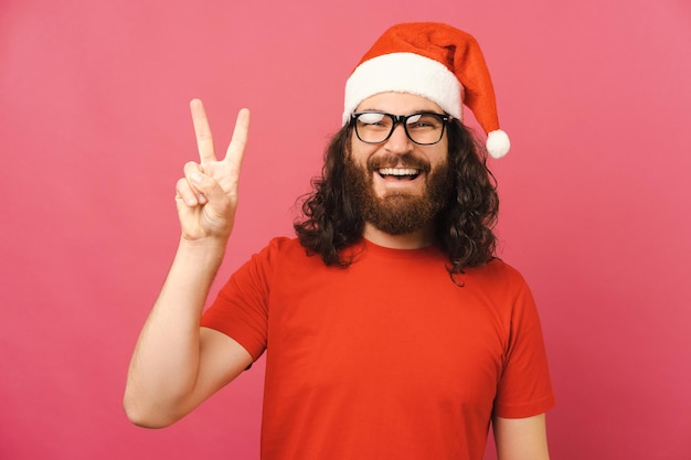 L'uomo barbuto estatico che indossa il cappello di Natale sta mostrando il gesto V mentre sorride