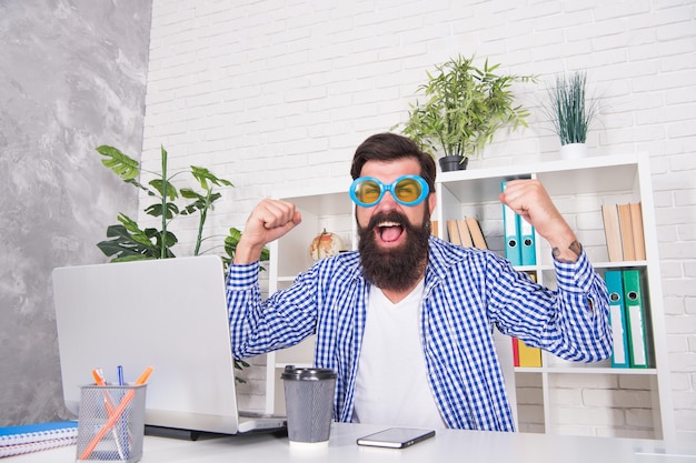 L'uomo barbuto felice celebra la vittoria facendo il gesto del vincitore al desktop in un ufficio moderno, celebrazione.