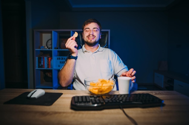 L'uomo barbuto felice si siede al lavoro al computer di notte e mangia patatine con un sorriso sul viso