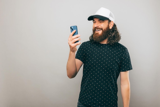 L'uomo barbuto felice sta mandando messaggi con qualcuno che sorride mentre si trova vicino a un muro grigio