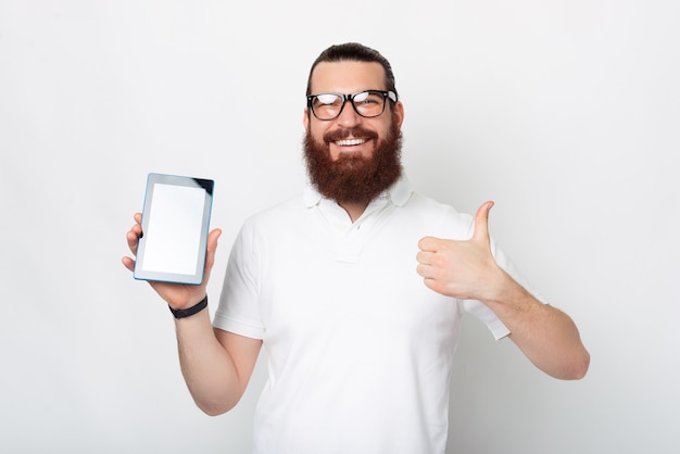 L'uomo barbuto felice sta mostrando il suo schermo del tablet alla fotocamera mentre mostra il gesto del pollice in su.