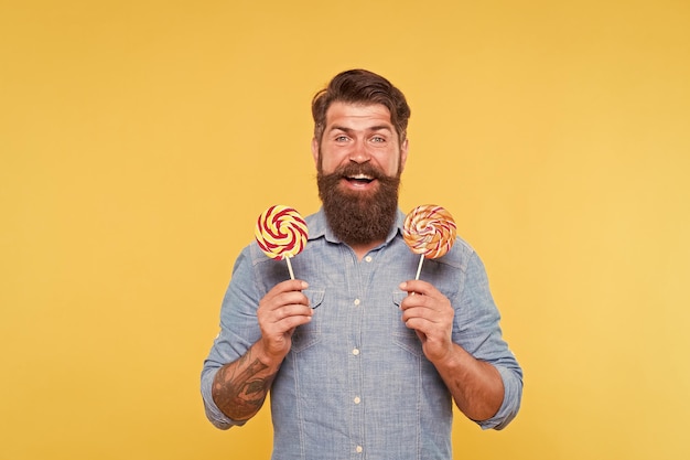 L'uomo barbuto felice tiene le gustose caramelle a vortice arcobaleno che si aprono ideali per la festa di compleanno e la celebrazione dello sfondo giallo del buffet di caramelle