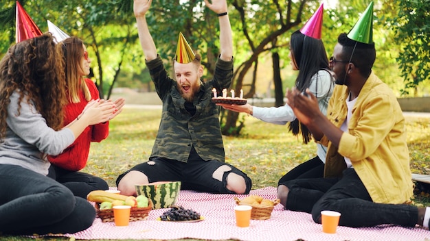 L'uomo barbuto festeggia il compleanno con gli amici nel parco che soffia le candeline sulla torta gridando ed esprimendo emozioni positive mentre gli amici ridono e battono le mani