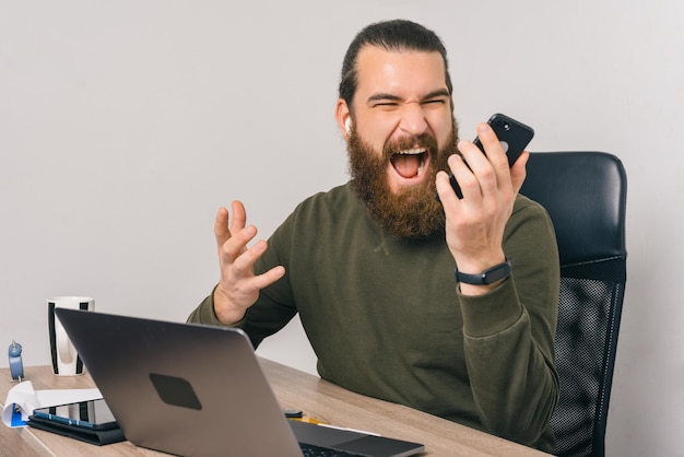 L'uomo barbuto frustrato sta gridando al telefono che tiene in mano.