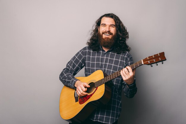 L'uomo barbuto gioioso tiene in mano e suona una chitarra su sfondo grigio.