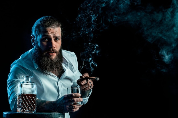 L'uomo barbuto hipster fuma un sigaro beve il whisky soffia il fumo vestito con una camicia bianca in studio