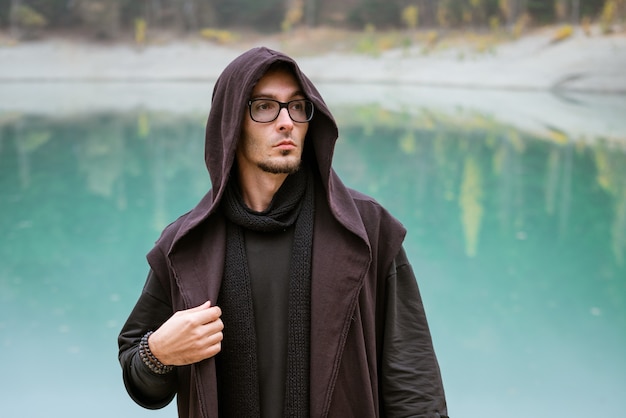 L'uomo barbuto in abiti casual con cappuccio e occhiali si trova sulla riva del lago turchese e guarda a s...