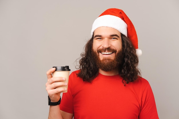 L'uomo barbuto riccio che indossa il cappello di Natale tiene in mano una tazza di caffè da asporto