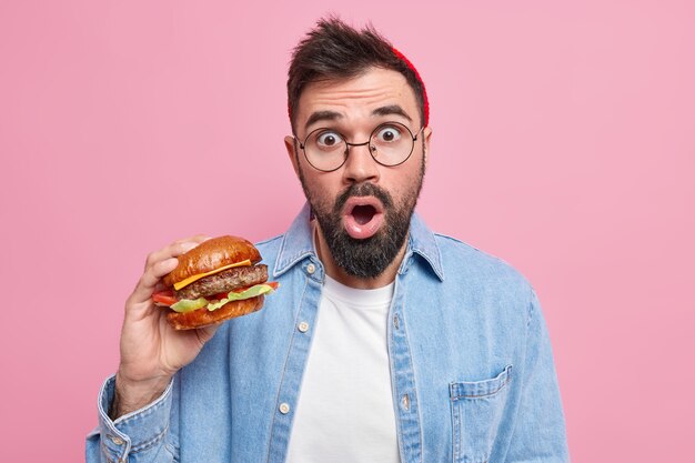L'uomo barbuto sbalordito mangia cibo ad alto contenuto calorico il delizioso hamburger gode del gusto di un hamburger fresco vestito con abiti casual casual