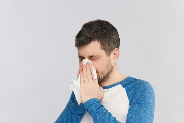 L'uomo barbuto soffia il naso nel tessuto con gli occhi chiusi che indossa una camicia con maniche blu isolate su grigio