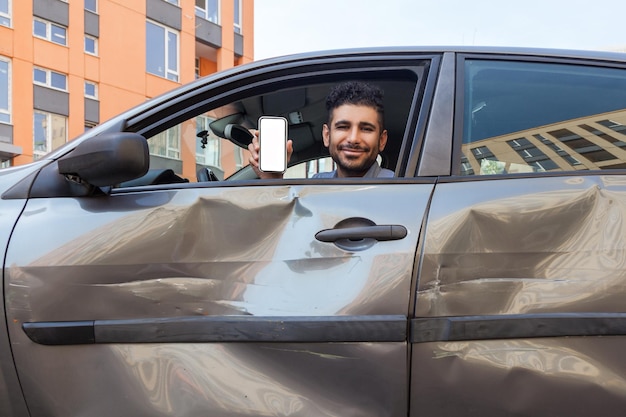 L'uomo barbuto sorridente positivo seduto in un'auto ammaccata e che mostra il telefono cellulare con lo schermo vuoto alla fotocamera offre servizi di ispezione assicurativa dopo un incidente stradale Scatto all'aperto