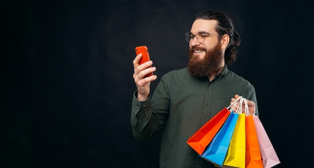 L'uomo barbuto sta guardando il telefono rosso e tiene in mano alcune borse della spesa Studio girato su sfondo nero