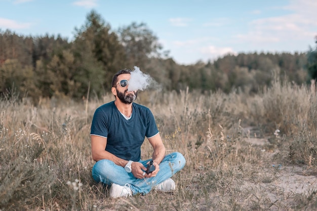 L'uomo barbuto sta riposando in una radura della foresta e sta svapando e sfogandosi da un'attività di svapo