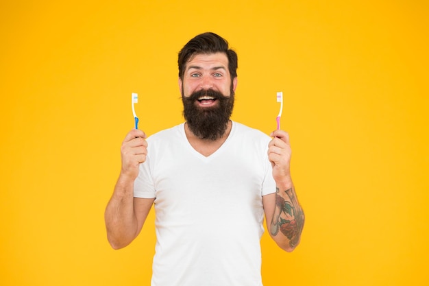 L'uomo barbuto tiene gli spazzolini da denti sfondo giallo Il ragazzo tiene due spazzolini da denti Hipster sorridente felice si prende cura dell'igiene Spazzola i denti concetto Igiene dei denti Tecnologia di pulizia dei denti ad ultrasuoni