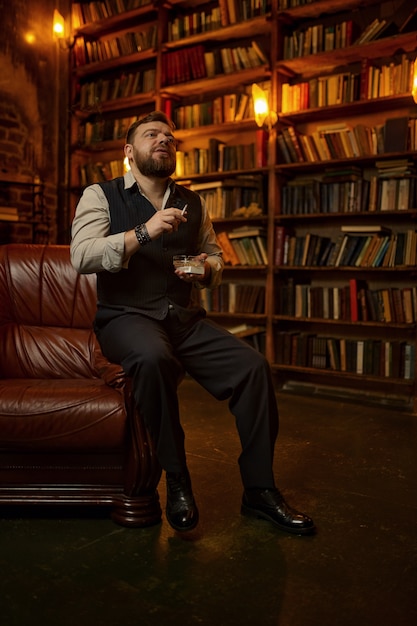 L'uomo barbuto tiene il portacenere e fuma la sigaretta, la libreria e l'interno dell'ufficio ricco. Cultura del fumo di tabacco, sapore specifico