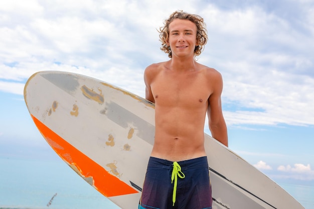 L'uomo bello cammina con la tavola da surf in bianco bianca aspetta l'onda per fare surf sulla riva dell'oceano del mare Concetto di sport fitness libertà felicità nuova vita moderna hipster