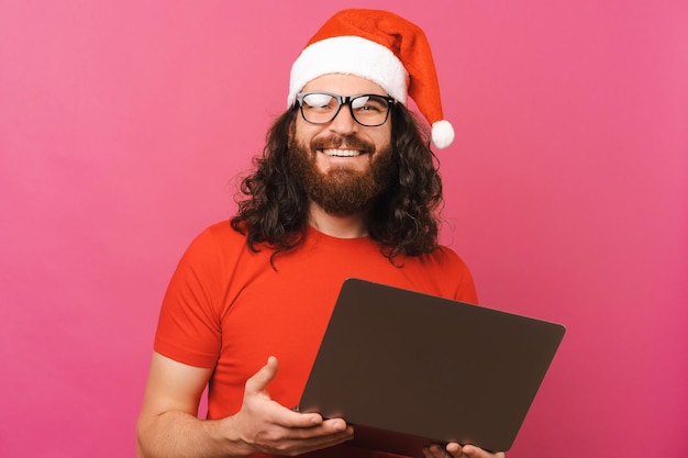 L'uomo bello con il cappello di Natale tiene in mano un laptop mentre sorride alla telecamera