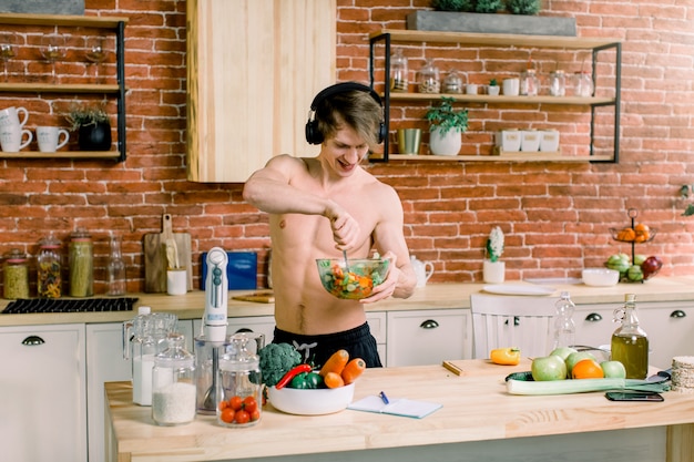 L'uomo bello felice ascolta musica in cuffie e balla, cucinando nella cucina a casa.