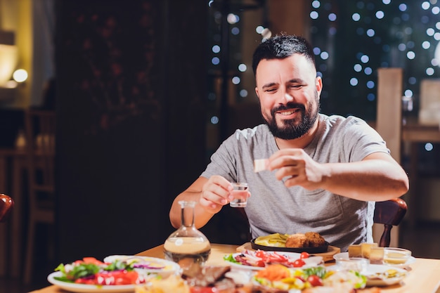 L'uomo beve la vodka a un grande tavolo con cibi e snack grassi.