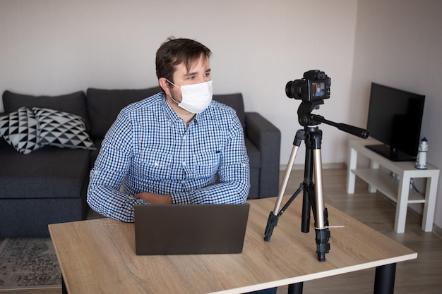 L'uomo blogger che indossa una maschera protettiva sta registrando video sulla videocamera. Blogger maschio in streaming sulla pandemia di coronavirus. Resta a casa per l'auto-quarantena di contenimento a distanza sociale