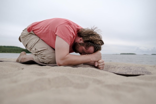 L'uomo calmo fa yoga, fa l'esercizio sul tappeto sulla riva sabbiosa del lago