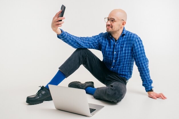 L'uomo caucasico bello senza capelli in camicia blu, pantaloni neri e calzini blu fa il libero professionista con il computer portatile e fa il selfie.