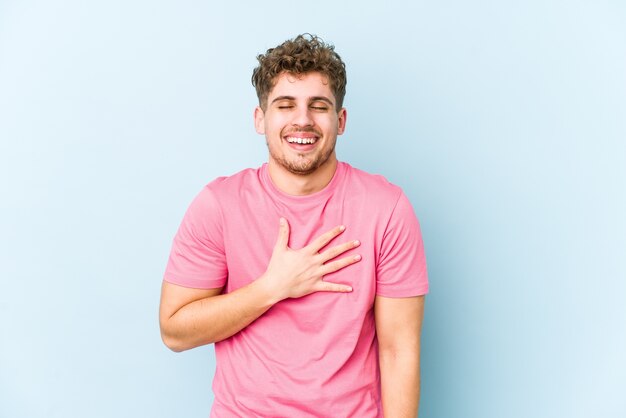 L'uomo caucasico dei giovani capelli ricci biondi isolato ride ad alta voce mantenendo la mano sul petto.