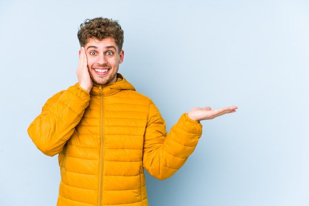 L'uomo caucasico dei giovani capelli ricci biondi tiene lo spaceon in bianco un palmo, tiene la mano sulla guancia.