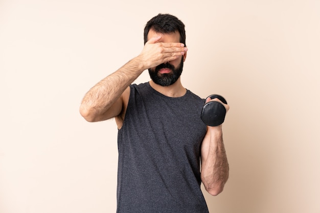 L'uomo caucasico di sport con la barba che fa il sollevamento pesi che copre gli occhi a mano. Non voglio vedere qualcosa