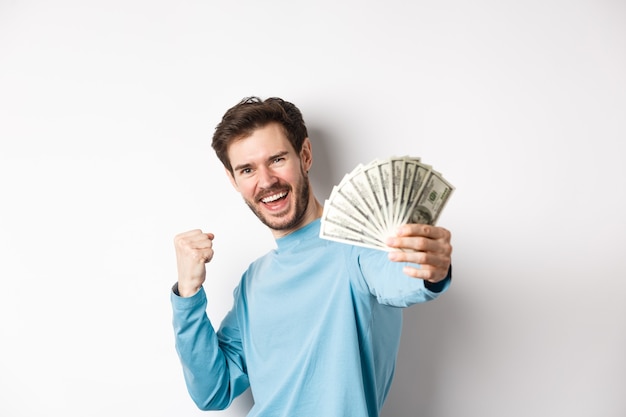 L'uomo caucasico felice allunga la mano con i soldi in dollari, dicendo sì e celebrando il reddito, ha ottenuto un premio in denaro, in piedi su sfondo bianco