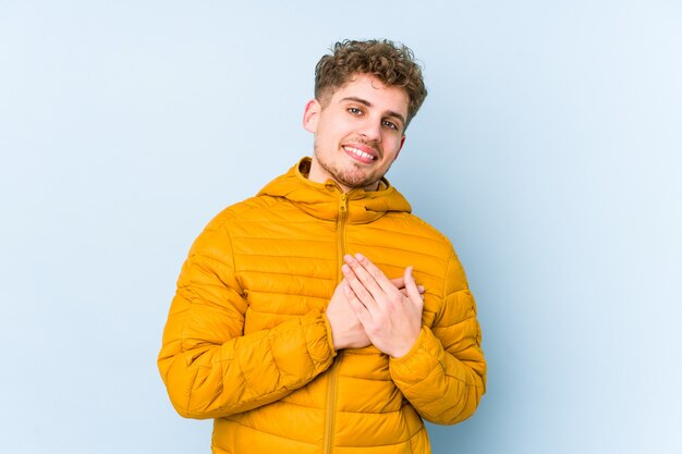 L'uomo caucasico giovane dei capelli ricci biondi isolato ha espressione amichevole, premendo il palmo al petto. Concetto di amore.