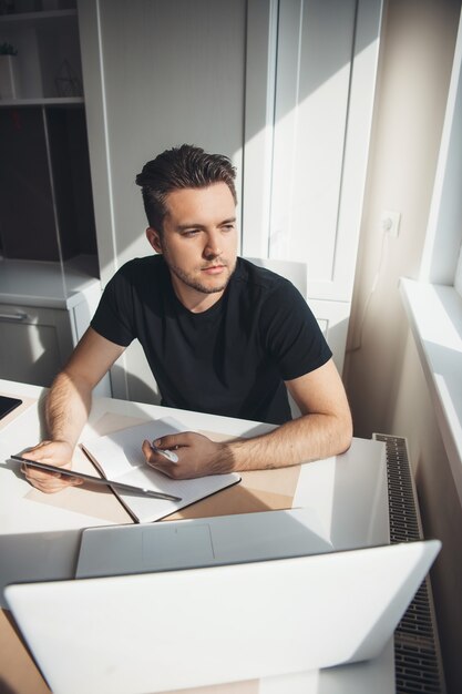 L'uomo caucasico sta lavorando a distanza da casa utilizzando un laptop e pensando a qualcosa vicino alla finestra