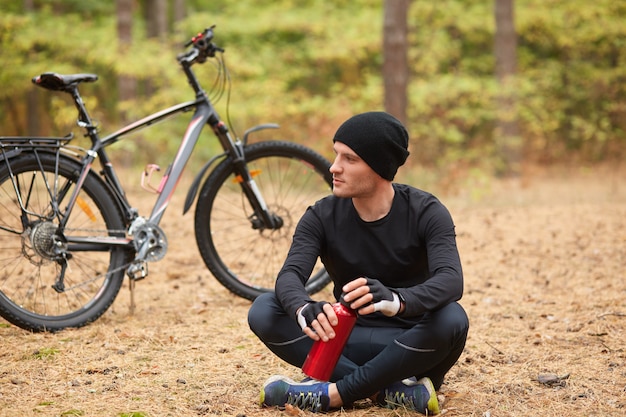 L'uomo che indossa tuta nera e cappuccio seduto onground con le gambe incrociate vicino alla sua bicicletta, il ciclista si ferma per bere acqua