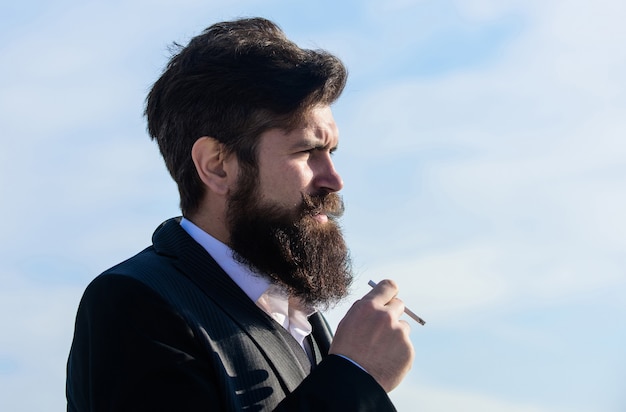 L'uomo con barba e baffi tiene la sigaretta. Fondo del cielo blu della sigaretta di fumo dei pantaloni a vita bassa barbuto. Il ragazzo con la sigaretta gode dell'influenza della nicotina. Verità sul piacere del fumo e sulla dipendenza da nicotina.