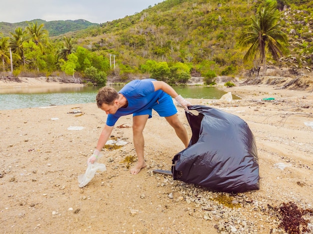 L'uomo con i guanti raccoglie i sacchetti di plastica che inquinano il mare Problema di spazzatura fuoriuscita di spazzatura sulla spiaggia di sabbia causata dall'inquinamento artificiale e dalla campagna ambientale per pulire il volontariato nel concetto