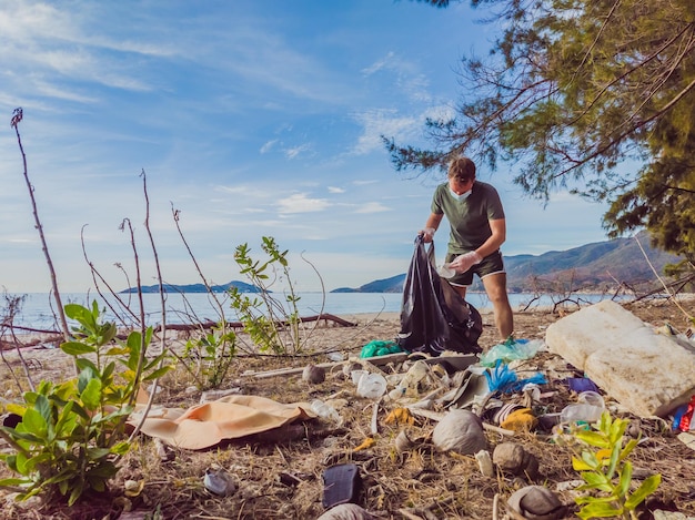 L'uomo con i guanti raccoglie la plastica che inquina il mare e la foresta Problema di spazzatura fuoriuscita di spazzatura sulla spiaggia di sabbia causata dalla campagna di inquinamento artificiale per pulire il volontario nel concetto