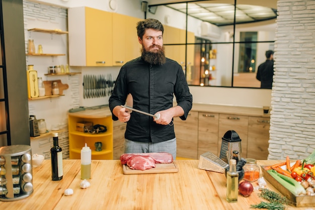 L'uomo con il coltello si prepara a tagliare la carne cruda. Cuoco unico che cucina il filetto con verdure, spezie ed erbe aromatiche